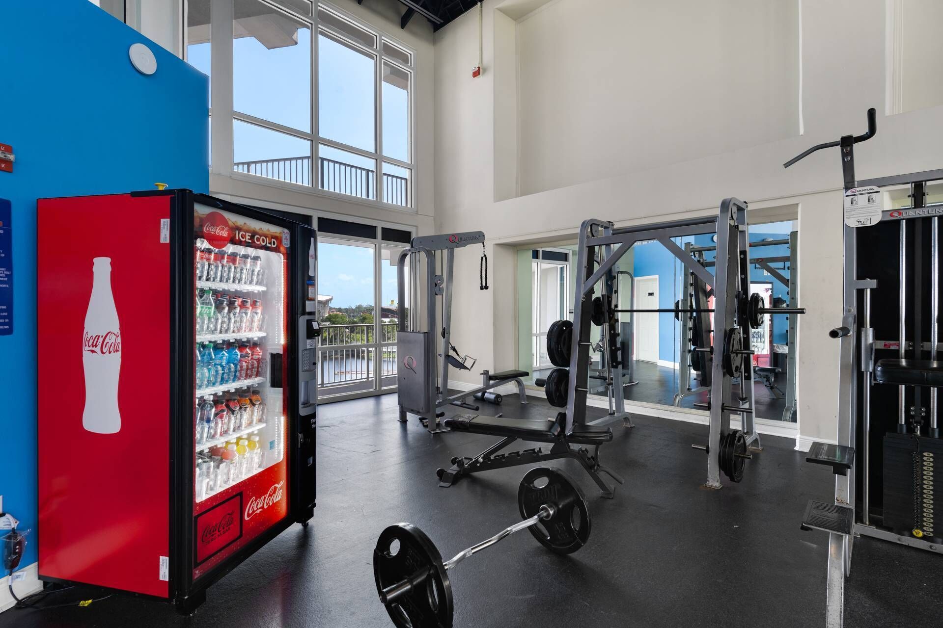 Une salle de sport équipée de matériel de musculation, d’un réfrigérateur à boissons Coca-Cola et de grandes fenêtres.