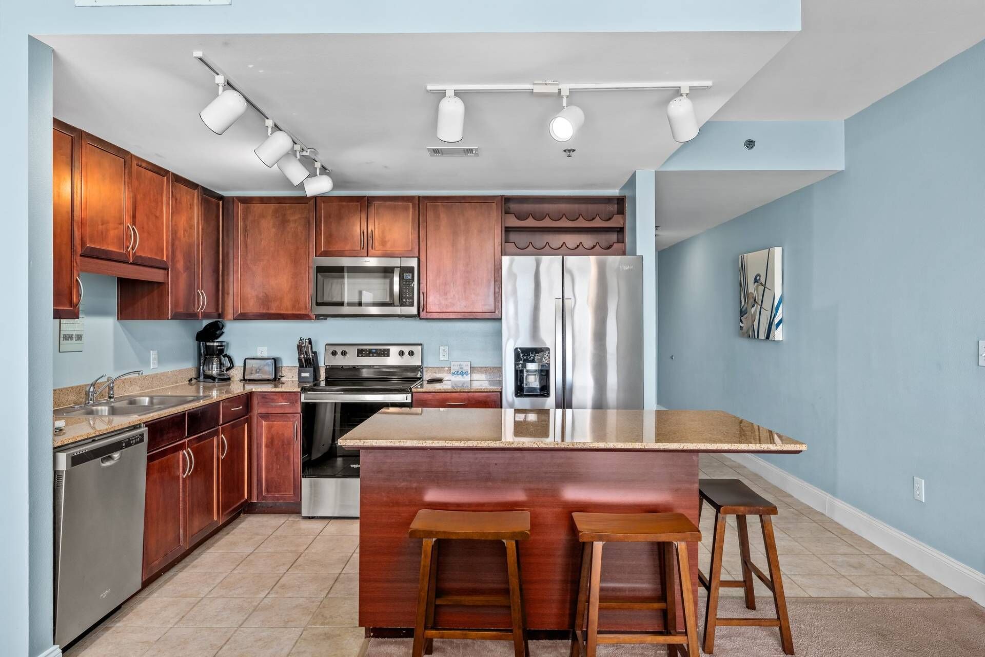 A modern kitchen with wooden cabinets, stainless steel appliances, a central island with stools, and a ceiling track lighting system.