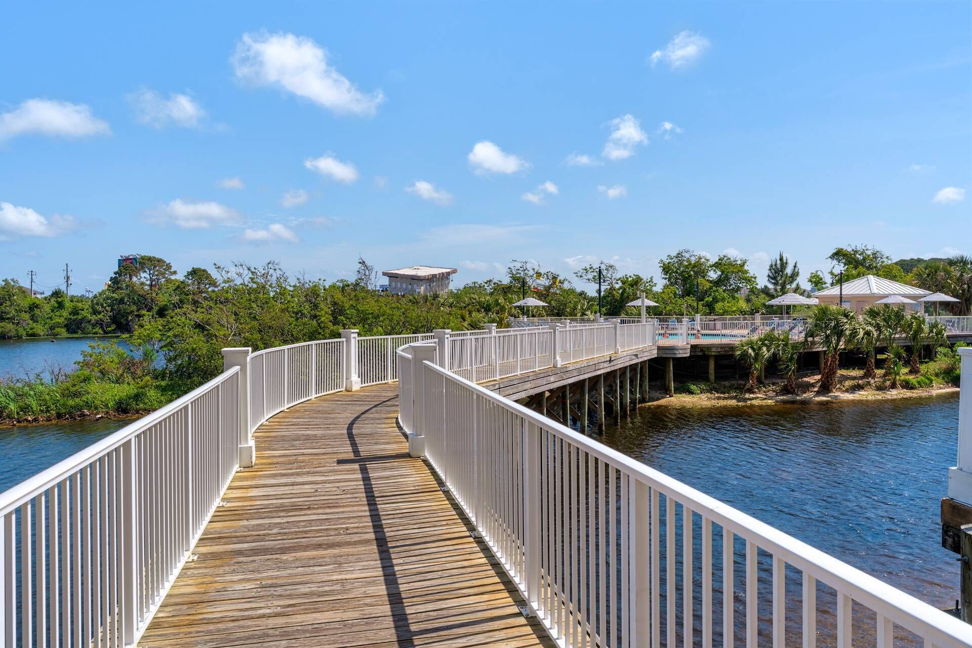 A wooden boardwalk leading to a pool area with white railings, surrounded by greenery and a clear blue sky.