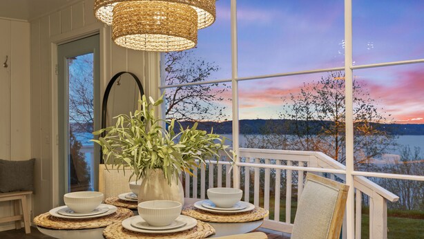 A dining area with a table set for four, a vase with greenery, and a view of a lake and trees at sunset.