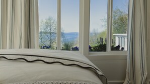 A bedroom with a large window, white curtains, and a view of trees and a distant landscape.