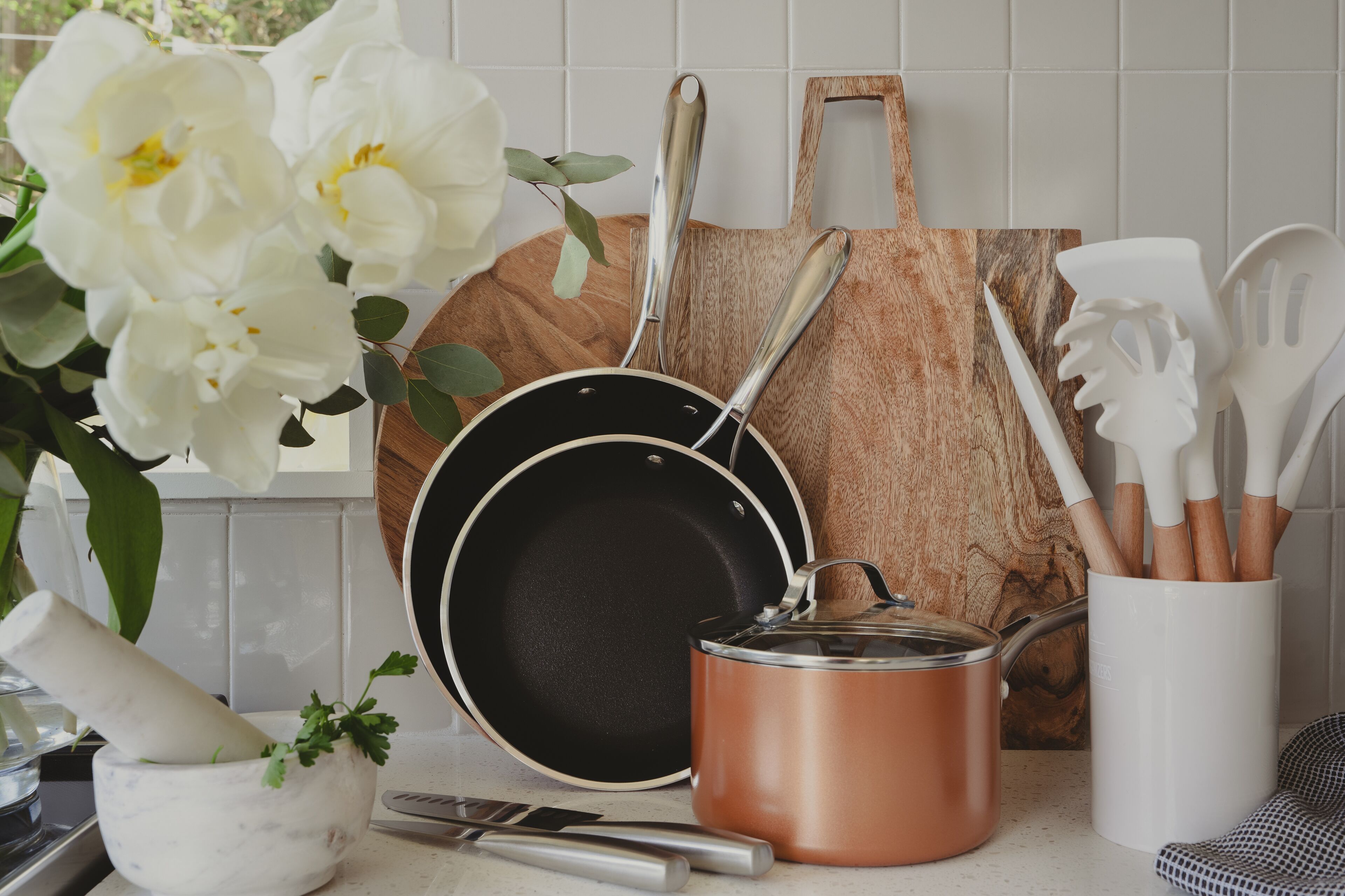 A kitchen counter with a frying pan, a copper pot, a wooden cutting board, and a set of wooden utensils.