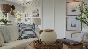 A cozy living room with a white sofa, striped pillows, a woven basket, and a large vase on a wooden table.