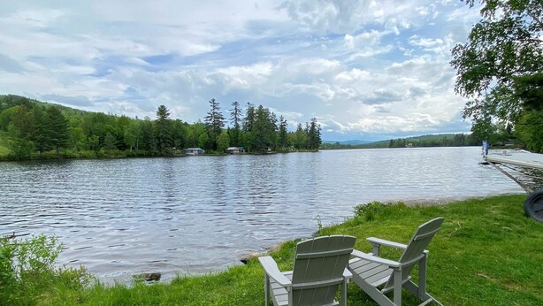 En vy över en sjö med två Adirondack-stolar placerade på en gräsyta, med utsikt över en stilla sjö och med träd och hus i bakgrunden.