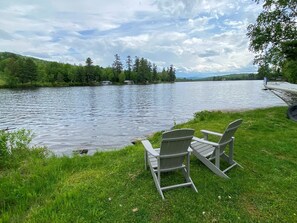 Vista lacustre con dos sillas Adirondack en un área verde, con vistas a un lago tranquilo con árboles y casas al fondo.