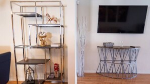 A living room with a metal shelving unit, a round metal table, and a flat-screen TV mounted on the wall. - NORTH HOLLYWOOD GEM: 2BR HOME w/ PRIVATE HOT TUB, PATIO/YARD & EV CHARGER p42 (Los Angeles)