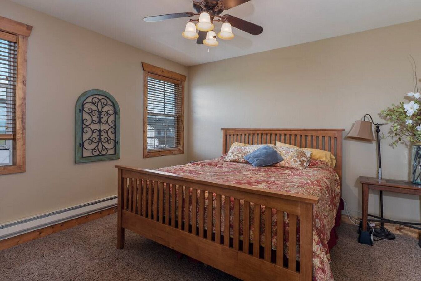 A bedroom with a wooden bed, two windows with blinds, a ceiling fan, and a bedside table with a lamp and vase.
