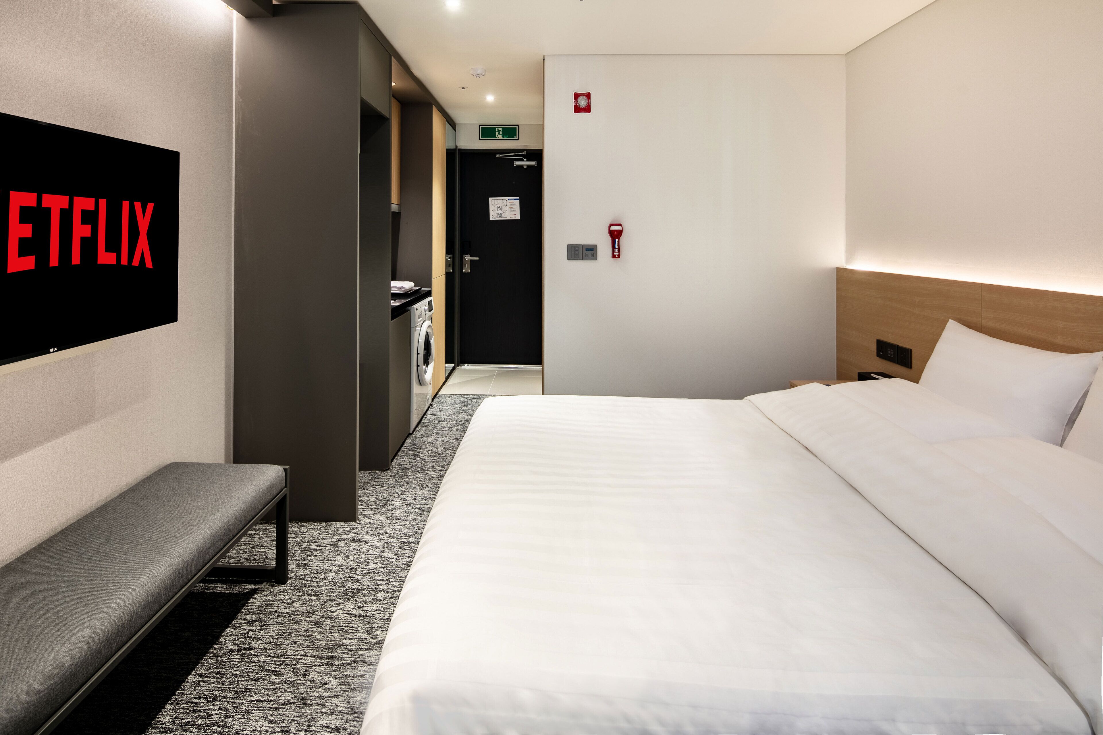 A hotel room with a bed, a bench, a wall-mounted TV displaying the Netflix logo, a door with a green exit sign, and a wall-mounted fire alarm.