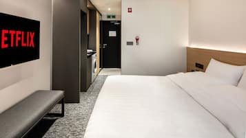 A hotel room with a bed, a bench, a wall-mounted TV displaying the Netflix logo, a door with a green exit sign, and a wall-mounted fire alarm.