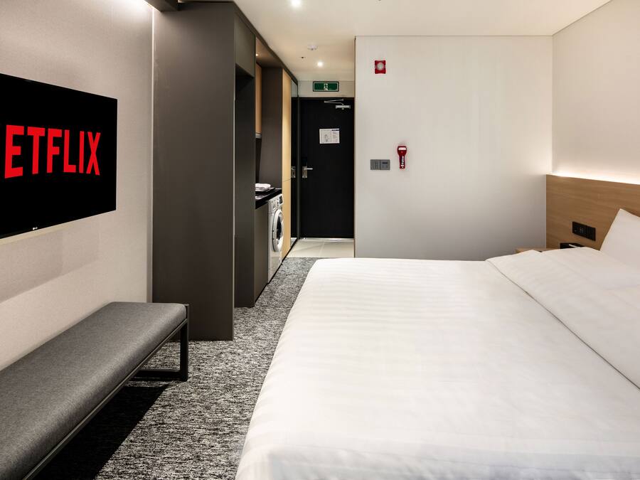 A hotel room with a bed, a bench, a wall-mounted TV displaying the Netflix logo, a door with a green exit sign, and a wall-mounted fire alarm.