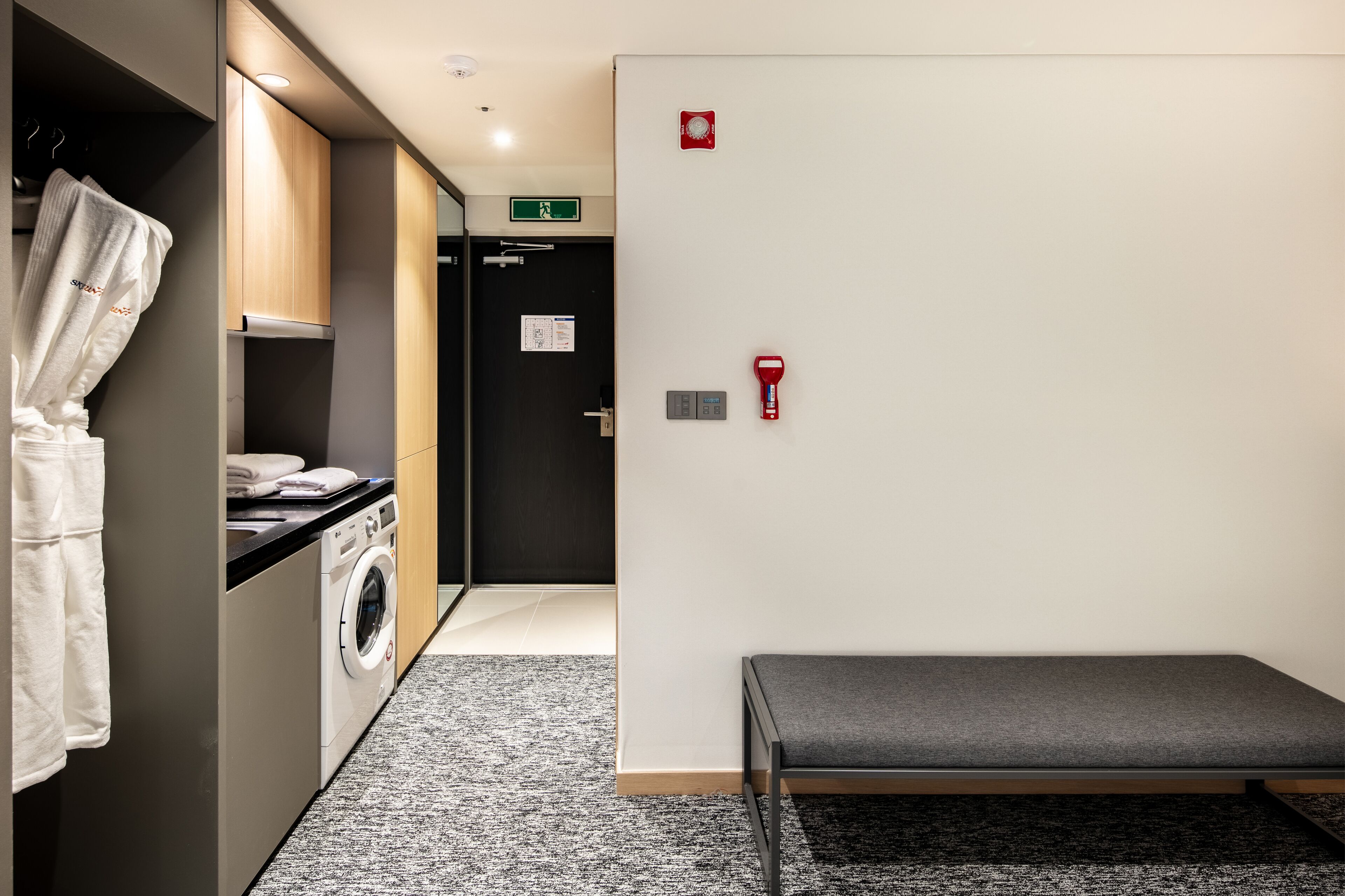 A hotel room with a washing machine, a bench, and a fire alarm.