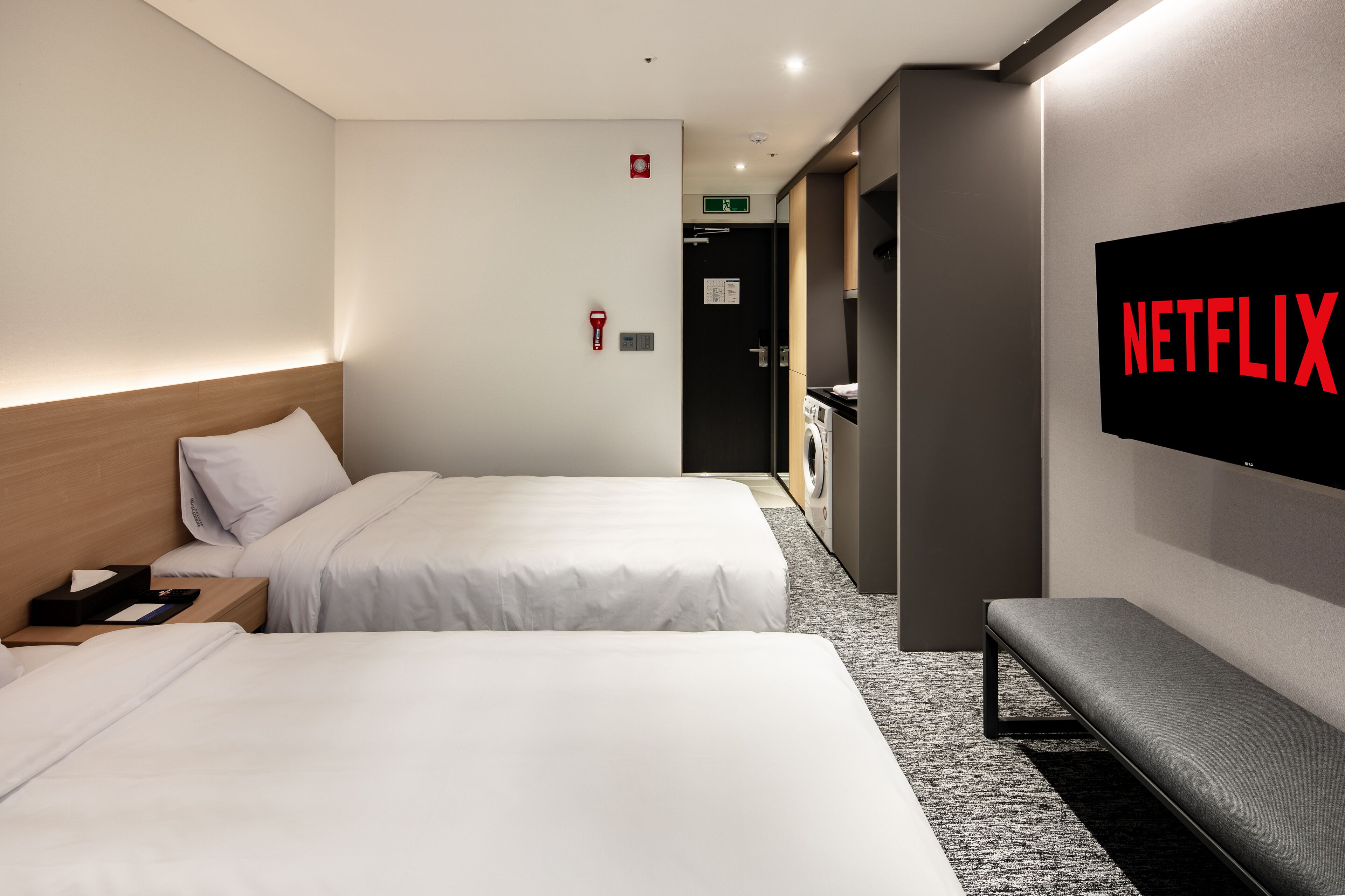 Une chambre d’hôtel avec deux lits, un banc, un écran de télévision affichant le logo « NETFLIX », et une machine à laver en arrière-plan.
