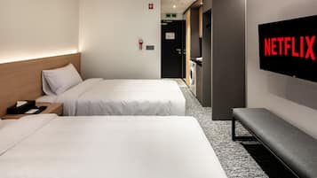 Une chambre d’hôtel avec deux lits, un banc, un écran de télévision affichant le logo « NETFLIX », et une machine à laver en arrière-plan.