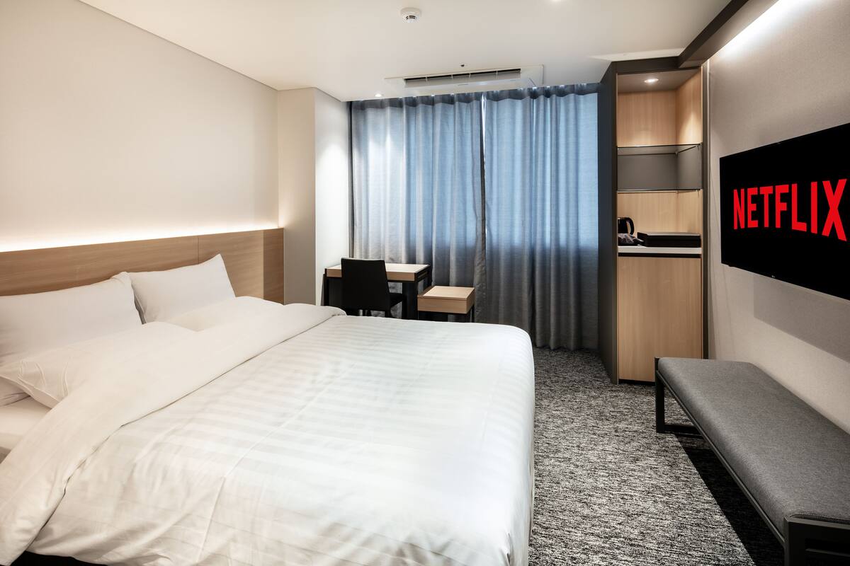 Habitación de hotel con cama, escritorio, banca, televisor y cortina.