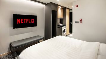 A modern hotel room with a large bed, a bench, a washing machine, and a television displaying "NETFLIX".