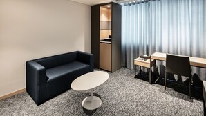 A modern hotel room with a dark blue sofa, a small round table, a wooden desk, and a large window with curtains. - Icheon Sky Sun Hotel (Icheon)