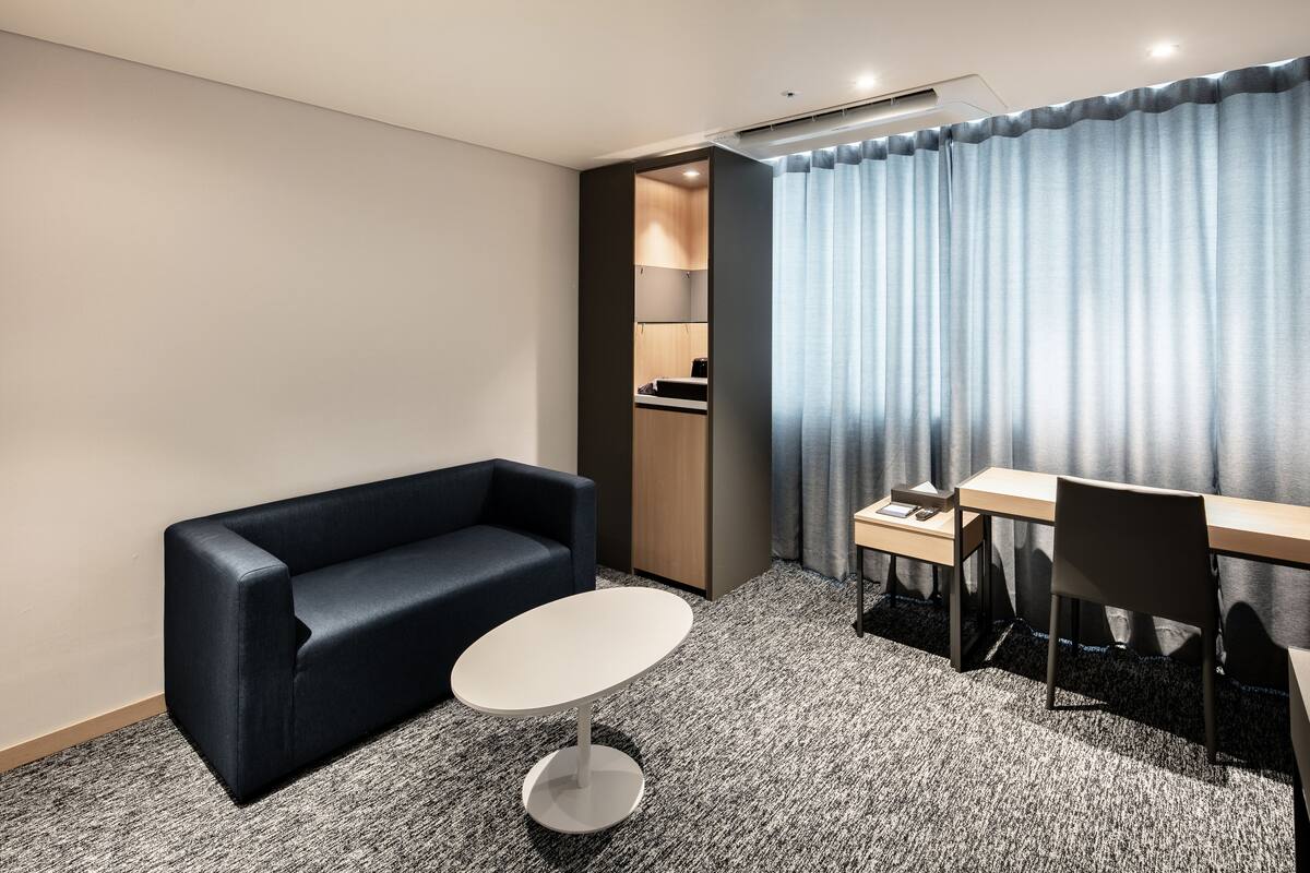 Una habitación de hotel moderna con un sofá azul oscuro, una pequeña mesa redonda, un escritorio de madera y un amplio ventanal con cortinas.