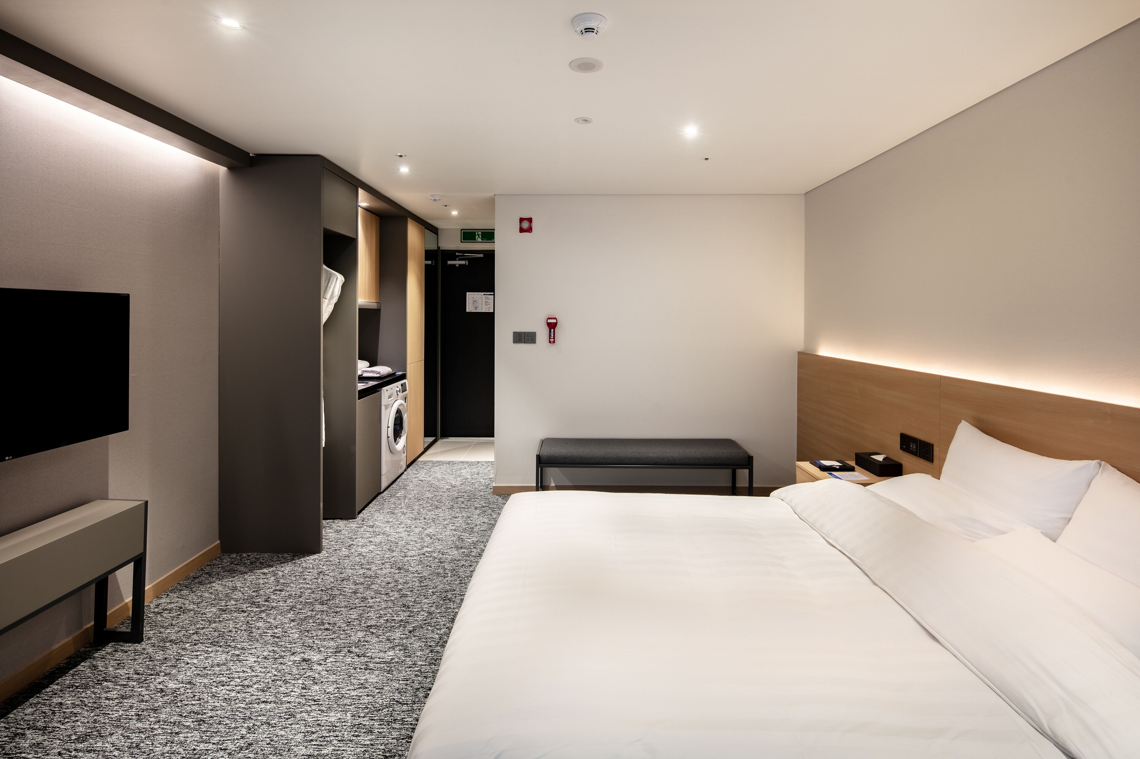 Quarto de hotel moderno com uma cama grande, televisão de tela plana, um banquinho, máquina de lavar e uma pequena escrivaninha com luminária.