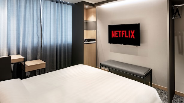 Habitación de hotel con cama, escritorio, banco, armario y televisor de pared mostrando el logotipo de Netflix.