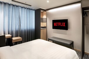Habitación de hotel con cama, escritorio, banco, armario y televisor de pared mostrando el logotipo de Netflix.