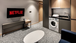 Una habitación de hotel moderna con una televisión de pantalla plana mostrando 'NETFLIX', una lavadora, un lavabo, una mesita pequeña y una silla azul.