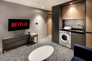 Una habitación de hotel moderna con una televisión de pantalla plana mostrando 'NETFLIX', una lavadora, un lavabo, una mesita pequeña y una silla azul.
