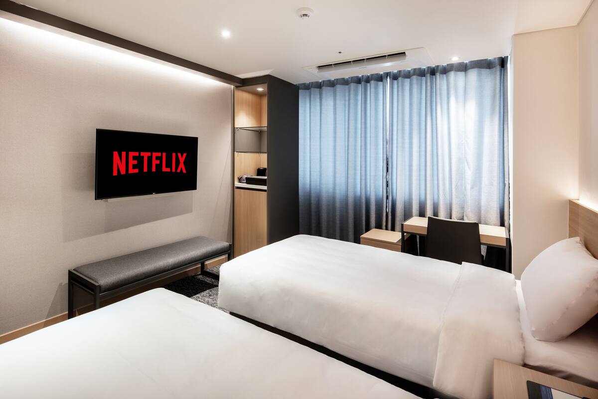 Habitación de hotel con dos camas, un televisor montado en la pared, un escritorio y una ventana con cortinas.