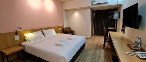 A hotel room with a bed, bedside table, chair, desk with a phone, water bottles, and a TV.