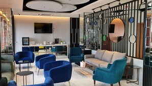 Een moderne woonkamer met blauwe fauteuils, een grijze bank en een salontafel. Er is een televisie aan de muur bevestigd en een decoratieve wand met spiegels en ronde elementen.