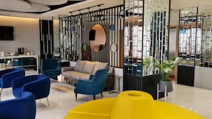Een moderne hotel lobby met blauwe en gele zitplaatsen, een flatscreen televisie en een decoratieve wand met spiegels en planten.