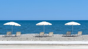 Tres sillas y sombrillas de playa en una arena pedregosa, con el océano al fondo.