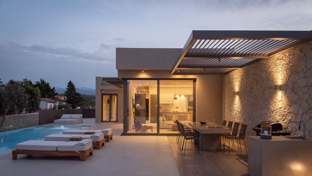 Un área de piscina moderna al aire libre con sillones de descanso, zona de comedor con mesa y sillas, y un muro de piedra con iluminación.