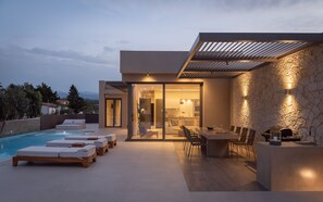 A modern outdoor pool area with lounge chairs, a dining area with a table and chairs, and a stone wall with lighting.