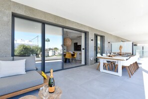 Un espace de vie extérieur moderne, comprenant un canapé, une table à manger et des chaises, une bouteille de champagne, et une vue sur le paysage urbain à travers de grandes portes vitrées.