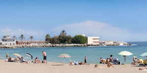 Eine Strandansicht mit badenden und sonnenden Personen, einem Felssteg und einem Gebäude im Hintergrund.
