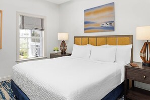 A neatly made bed with a white comforter and pillows, a wooden headboard, a nightstand with a lamp, and a window with a view of a balcony.