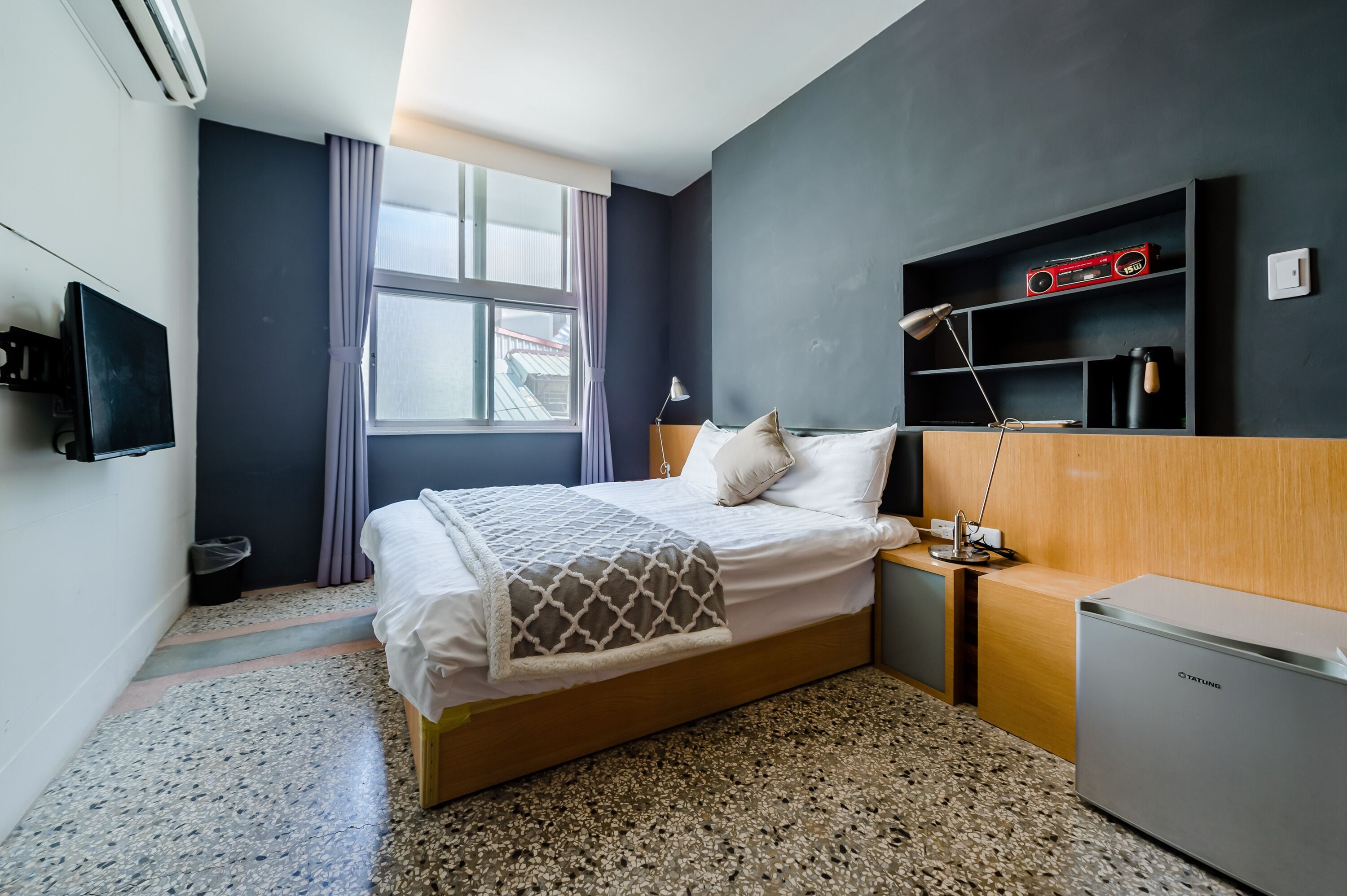 Una habitación de hotel moderna con cama, mesita de noche, lámpara, televisor y un refrigerador pequeño.