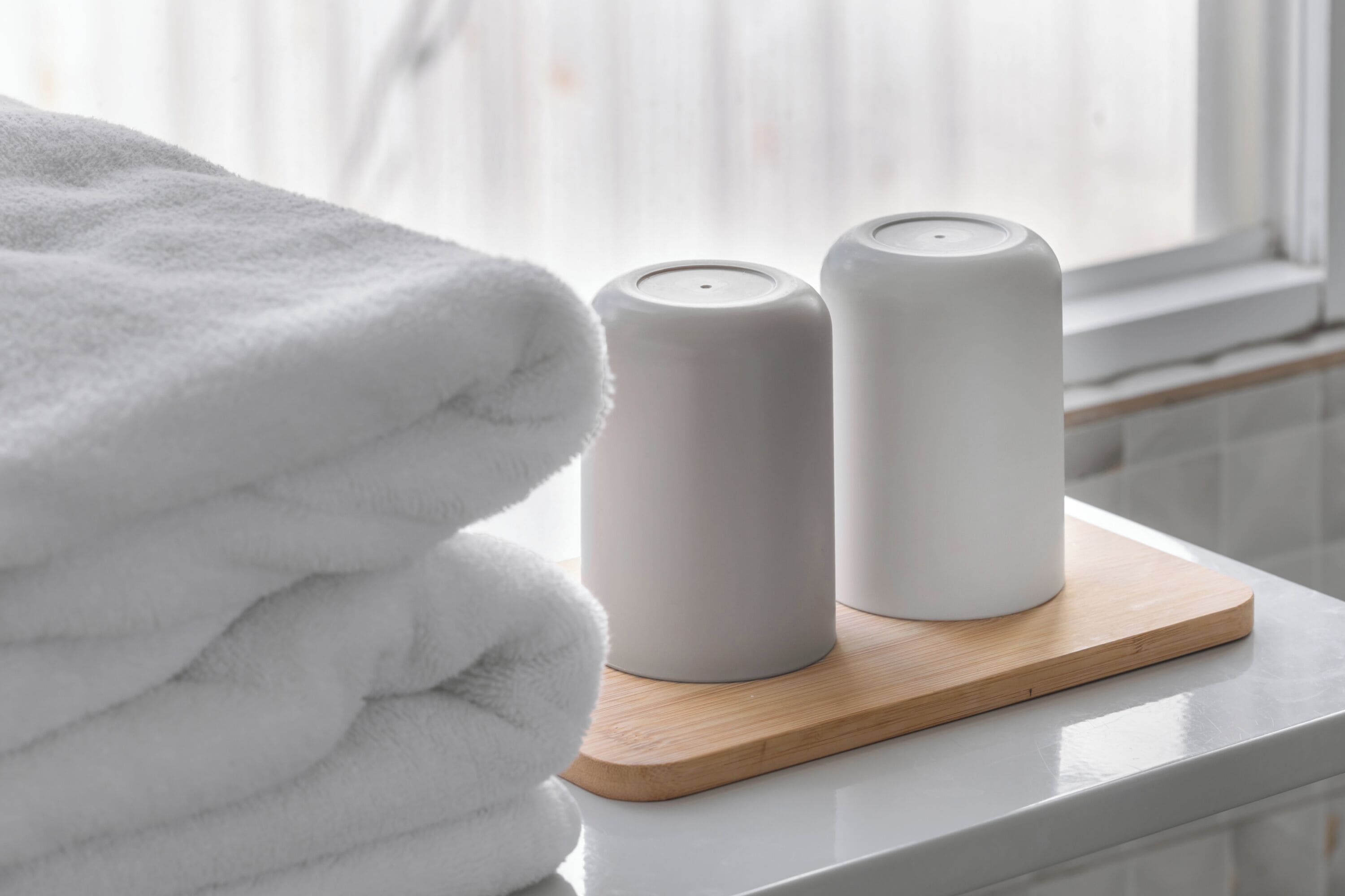 A neatly folded white towel on a wooden tray next to two white candles on a white surface.