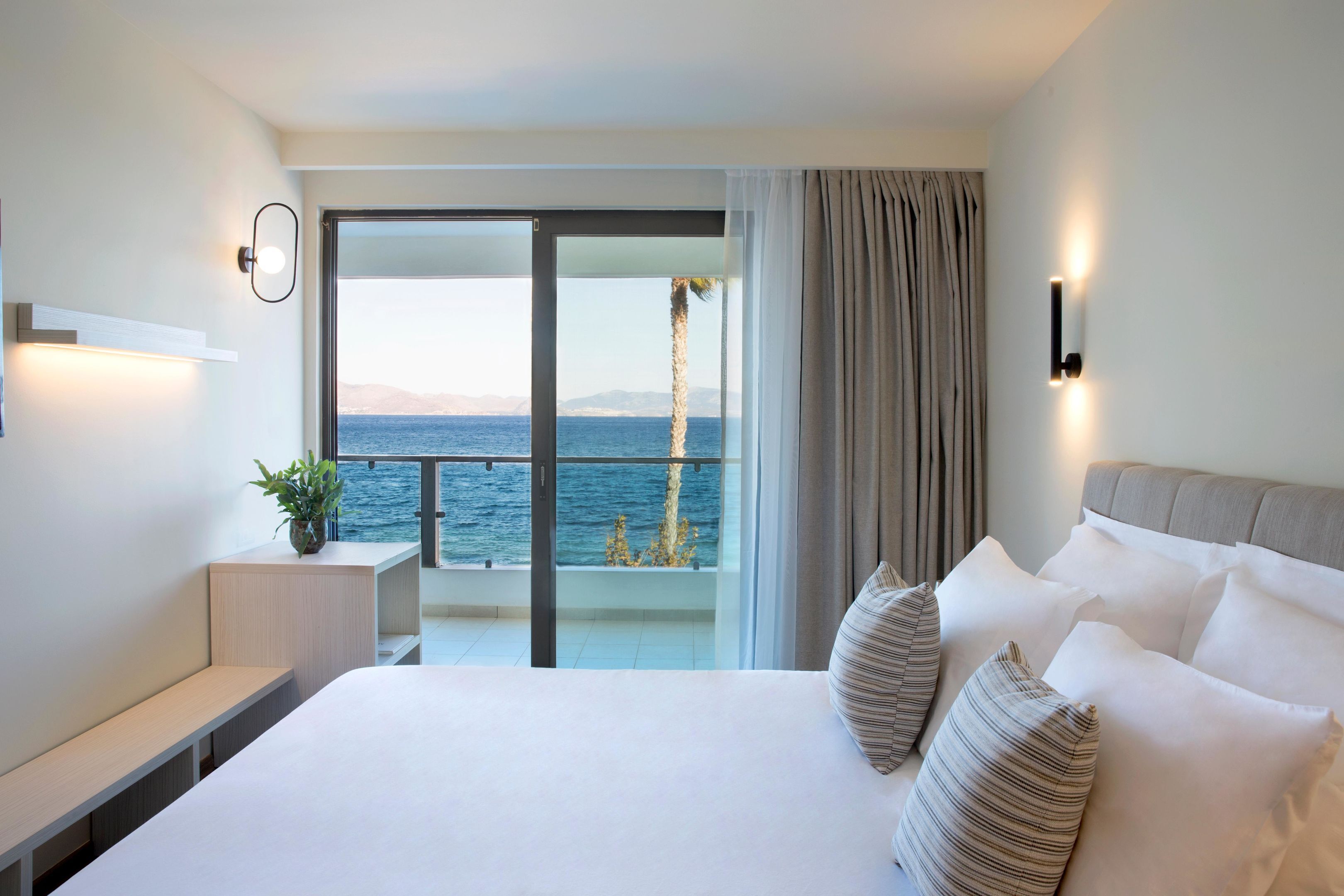Una camera d'albergo con un letto grande, un comodino, una pianta in vaso e vista sul mare attraverso una porta scorrevole in vetro.