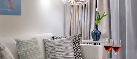 A modern living room with a sofa, patterned cushions, a round table with two glasses of red wine, a blue vase with a plant, and a hanging light fixture.
