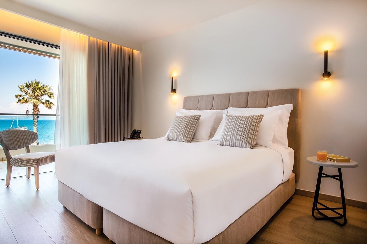A large bed with a wooden headboard, two bedside lamps, a chair, a small table with a book and a glass of orange juice, and a view of the ocean through a window with curtains.