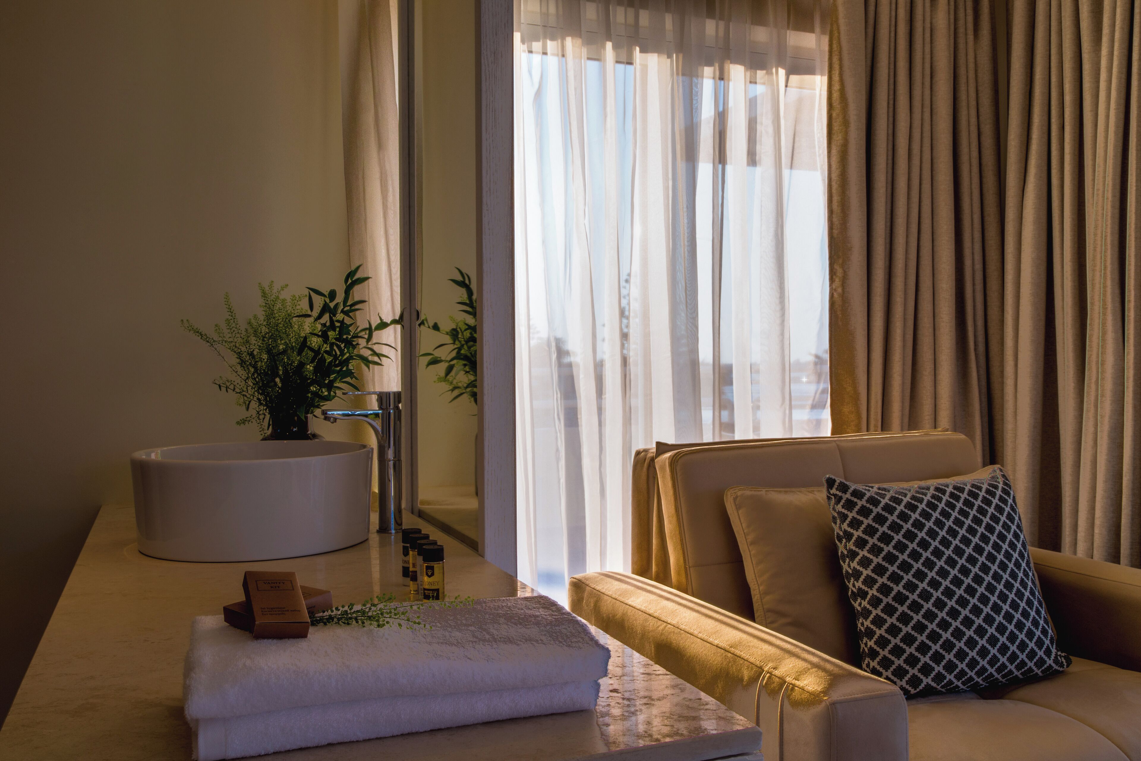 A modern hotel room with a sofa, a side table with toiletries, a potted plant, and a view of the outdoors through sheer curtains.