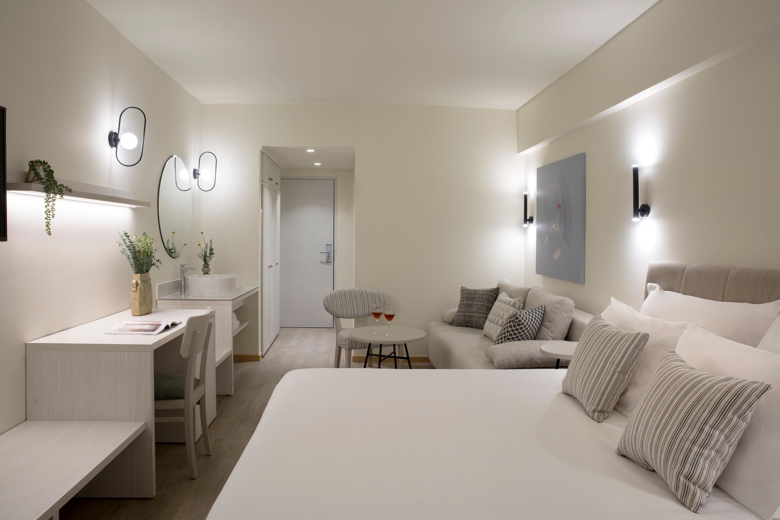 A modern hotel room with a large bed, a desk with a chair, a small round table, and a sofa.