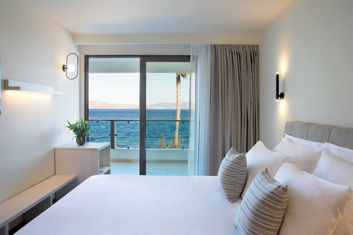 Een moderne hotelkamer met een groot bed, een nachtkastje en uitzicht op zee door een schuifglazen deur.