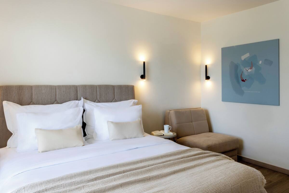 Een hotelkamer met een groot bed, twee wandlampen, een kleine tafel met een boek en een kopje, en een stoel.
