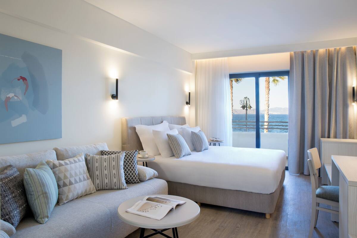 Een hotelkamer met een bed, een bank, een kleintje tafeltje met een boek en uitzicht op zee.