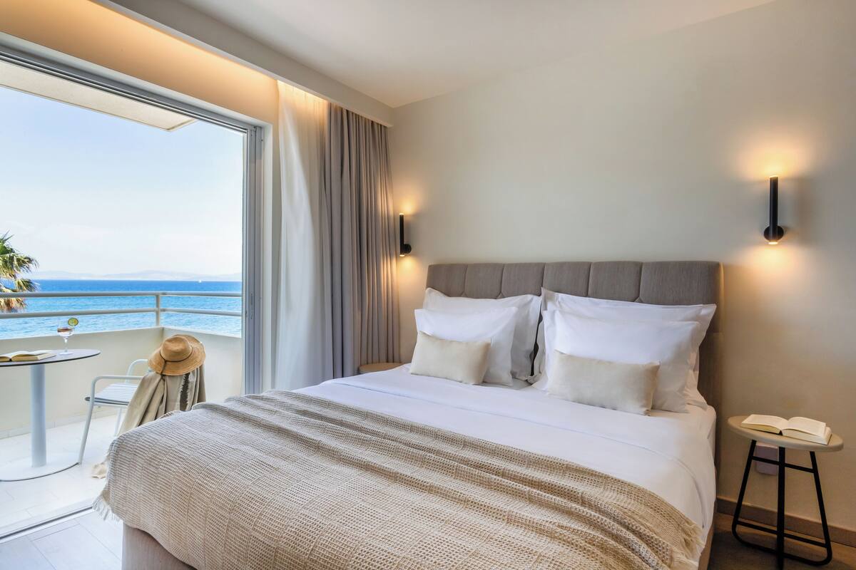 Hotelkamer met een groot bed, een balkon met uitzicht op zee en een tafeltje met een hoed en boek.
