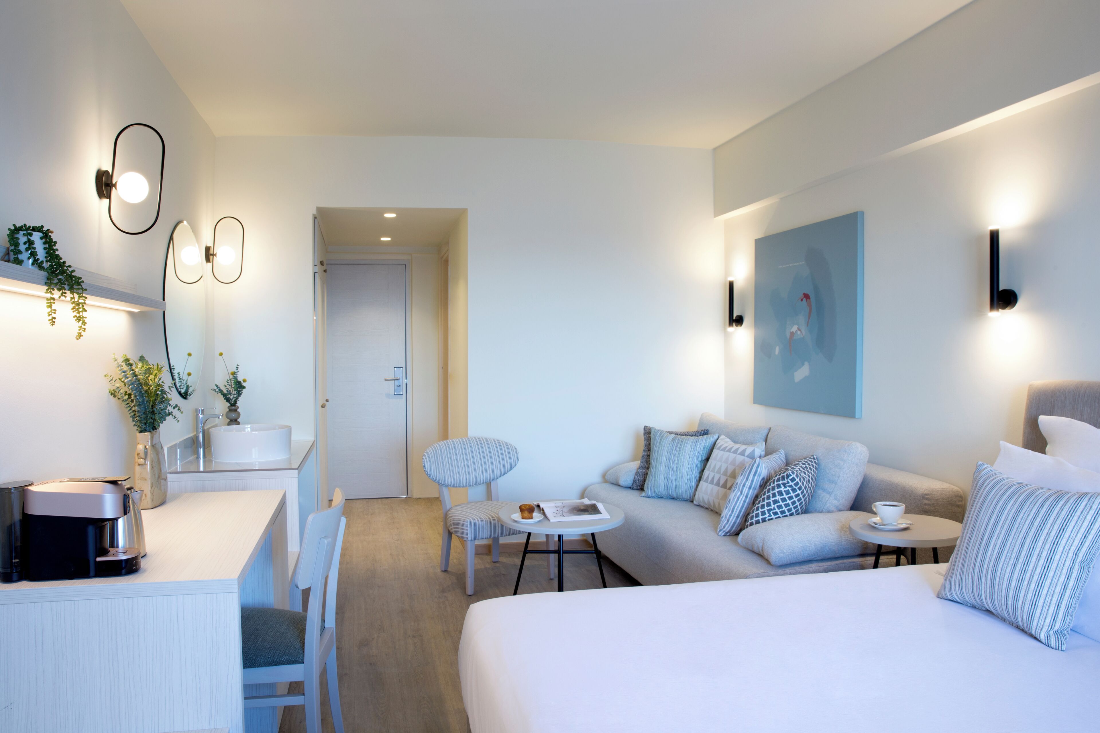 A modern hotel room with a bed, a sofa, a dining table, and a small kitchen area.