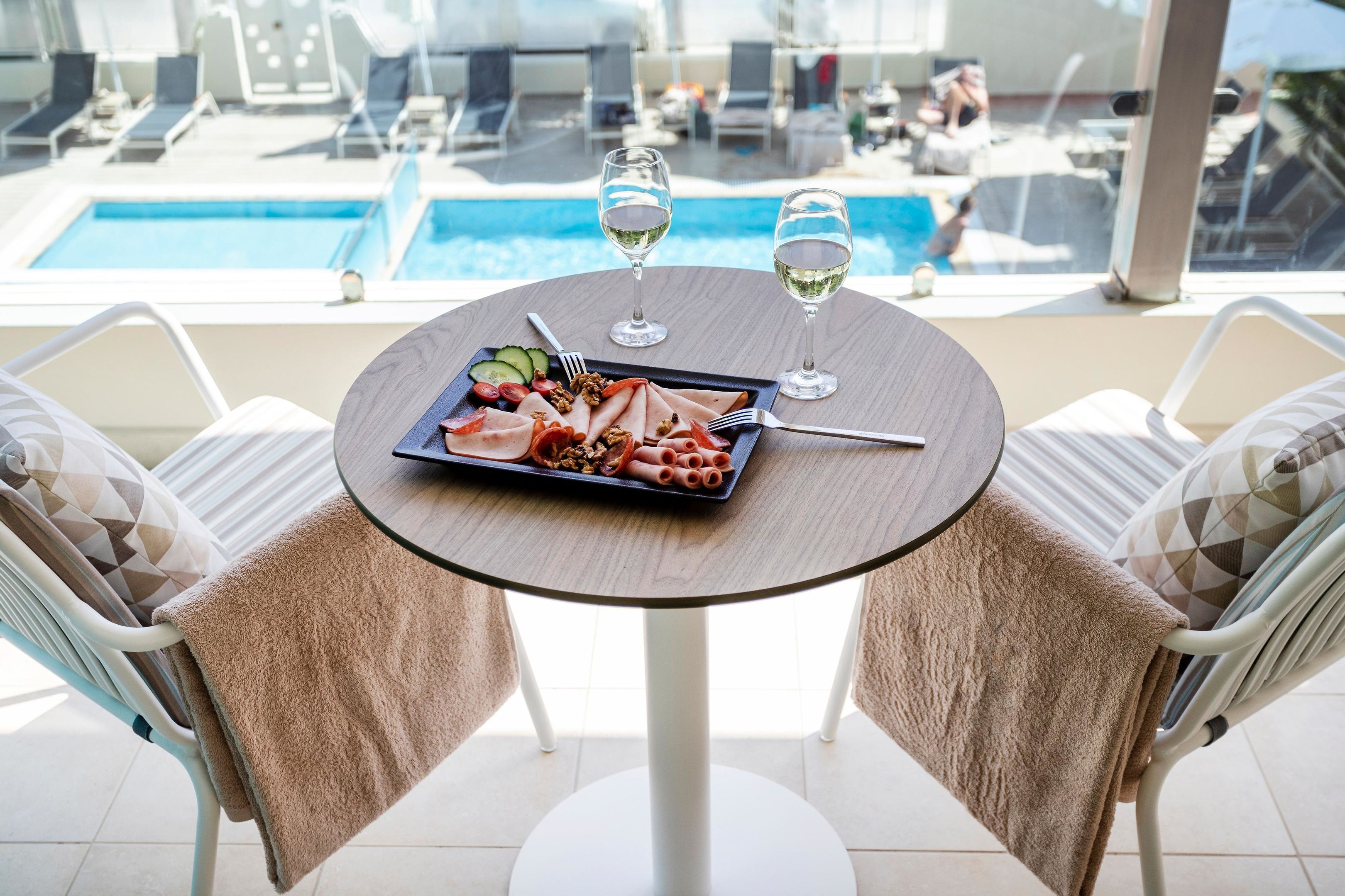 A dining table with a plate of food and two wine glasses, overlooking a pool area with lounge chairs.