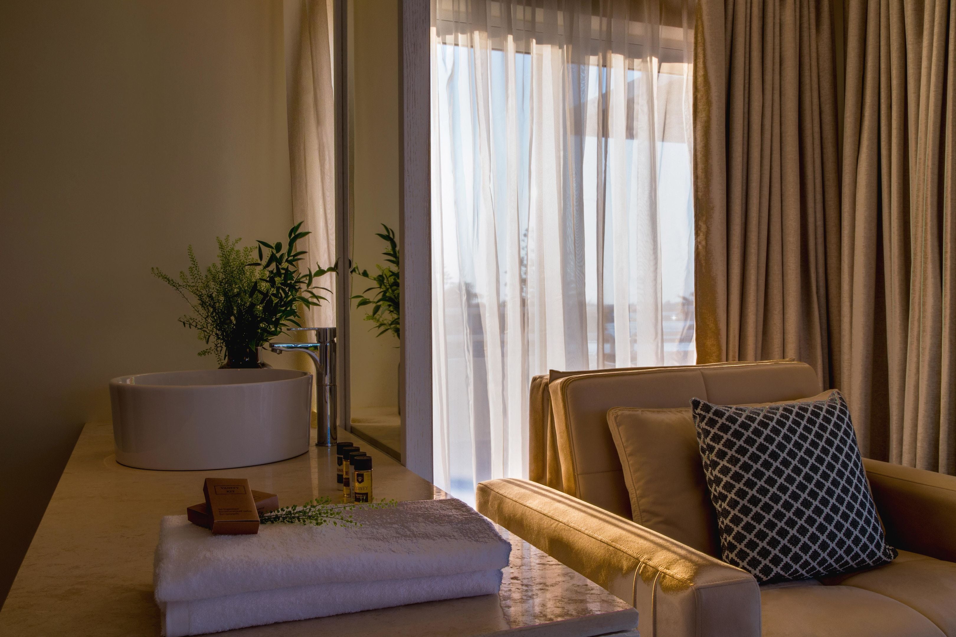 A modern hotel room with a sofa, a round white side table, a potted plant, and a window with sheer curtains.